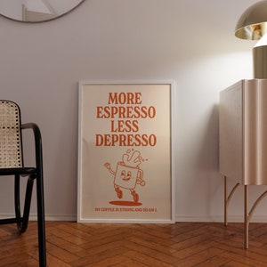 May include: A framed art print with the text "MORE ESPRESSO LESS DEPRESSO" in orange. The print features a cartoon coffee cup character with a smiling face and a splash of coffee. The text "MY COFFEE IS STRONG AND SO AM I" is at the bottom.