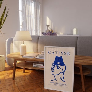 May include: A white framed poster with a blue illustration of a person with a cat on their head. The poster has the text "CATISSE" and "BERGGRUEN & CIE" in black.