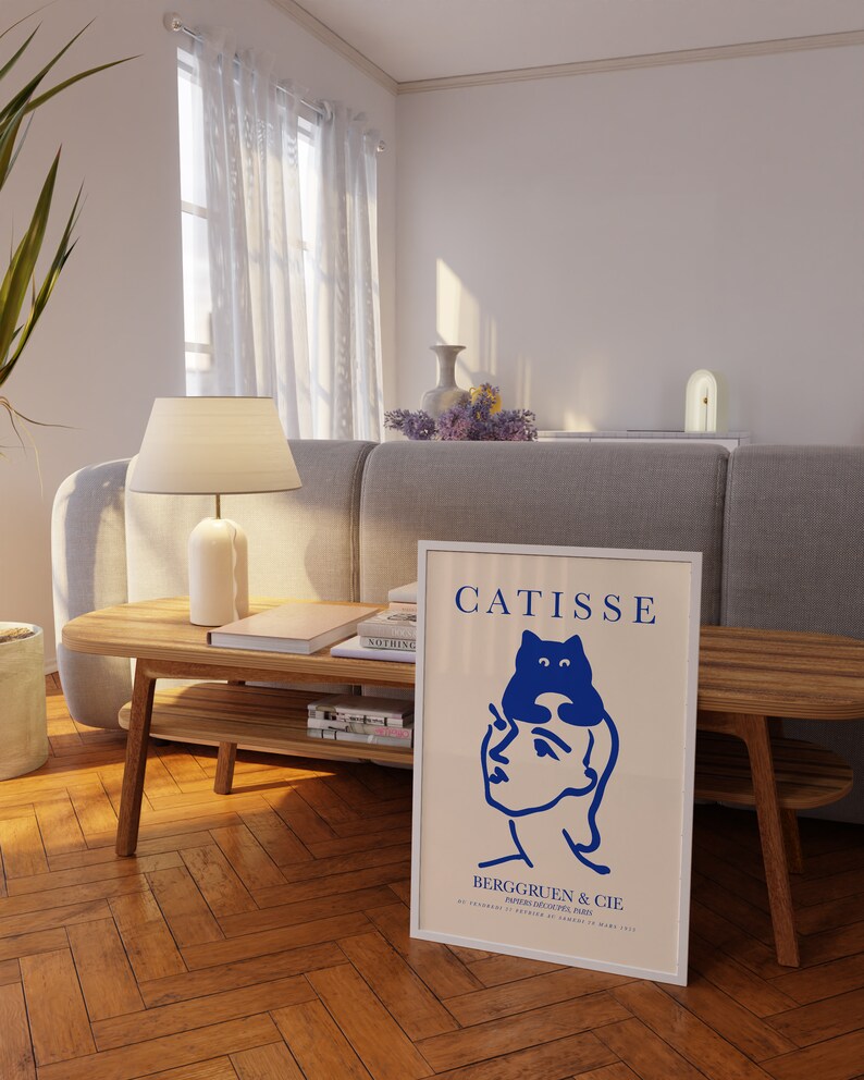 May include: A white framed poster with a blue illustration of a person with a cat on their head. The poster has the text "CATISSE" and "BERGGRUEN & CIE" in black.