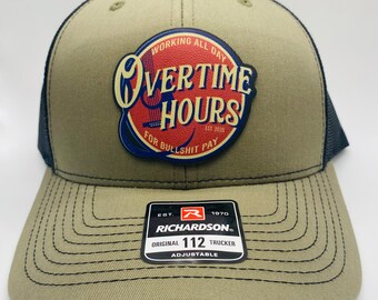 May include: A green and black trucker hat with a patch that says "Overtime Hours" in red and blue lettering. The patch also says "Working All Day" and "For Bullshit Pay". The hat is adjustable and has a Richardson 112 label.