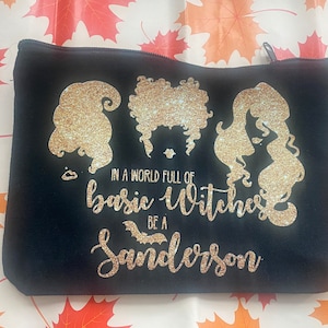 Könnte beinhalten: Schwarze Kosmetiktasche mit Reißverschluss, verziert mit goldenen Glitzer-Silhouetten von drei Hexen und dem Text "IN A WORLD FULL OF basic WITCHES BE A Sanderson". Geeignet für Make-up oder Toilettenartikel.