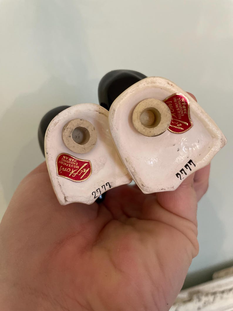 May include: A pair of white ceramic salt and pepper shakers shaped like a couple. The shakers have black painted details and red labels that say "Lady Gay" and "2777".