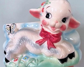 May include: A ceramic planter shaped like a lamb with a pink body, gray fur, black hooves, and a red bow. The lamb is lying on a blue and green cloud with a butterfly on the side.