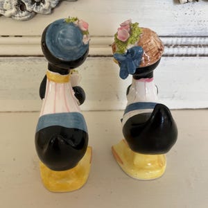 May include: A pair of vintage salt and pepper shakers shaped like ducks. The ducks are wearing blue and white outfits with floral hats. The shakers are made of ceramic and have a yellow base.