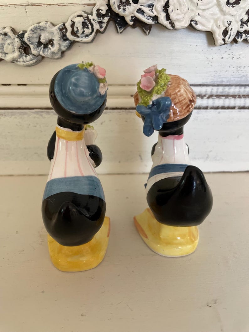 May include: A pair of vintage salt and pepper shakers shaped like ducks. The ducks are wearing blue and white outfits with floral hats. The shakers are made of ceramic and have a yellow base.