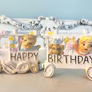 May include: Two ceramic birthday train cars with the words "HAPPY" and "BIRTHDAY". Each car features a smiling child and a dog, with colorful floral accents. The cars are painted in white, with gold and pastel details.