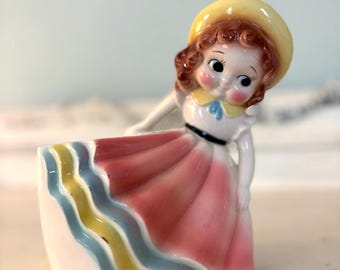 May include: A vintage ceramic figurine of a girl in a yellow hat and pink dress. The dress has a white base with wavy yellow, blue, and green stripes. The figurine has a cheerful expression and is likely a planter.