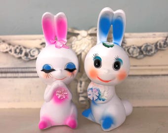 May include: Two vintage-style bunny figurines. One bunny has pink ears, a pink flower, and a pink flower on its chest. The other bunny has blue ears and a bouquet of flowers. Both bunnies have rosy cheeks, long eyelashes, and are white.