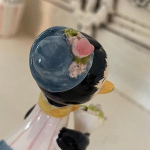 May include: A ceramic figurine of a woman with black hair wearing a blue hat with pink flowers. She is wearing a white and pink dress with a yellow sash. She is holding a bouquet of flowers.