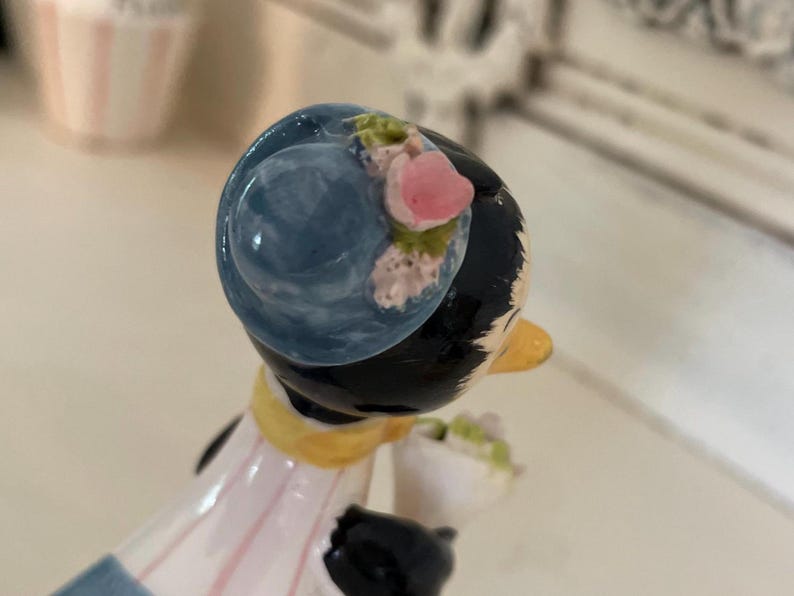 May include: A ceramic figurine of a woman with black hair wearing a blue hat with pink flowers. She is wearing a white and pink dress with a yellow sash. She is holding a bouquet of flowers.