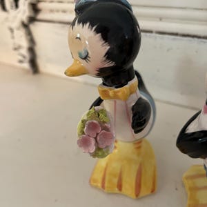 May include: A pair of ceramic penguin salt and pepper shakers. The female penguin is wearing a blue hat and holding a bouquet of pink flowers. The male penguin is wearing a black bow tie.