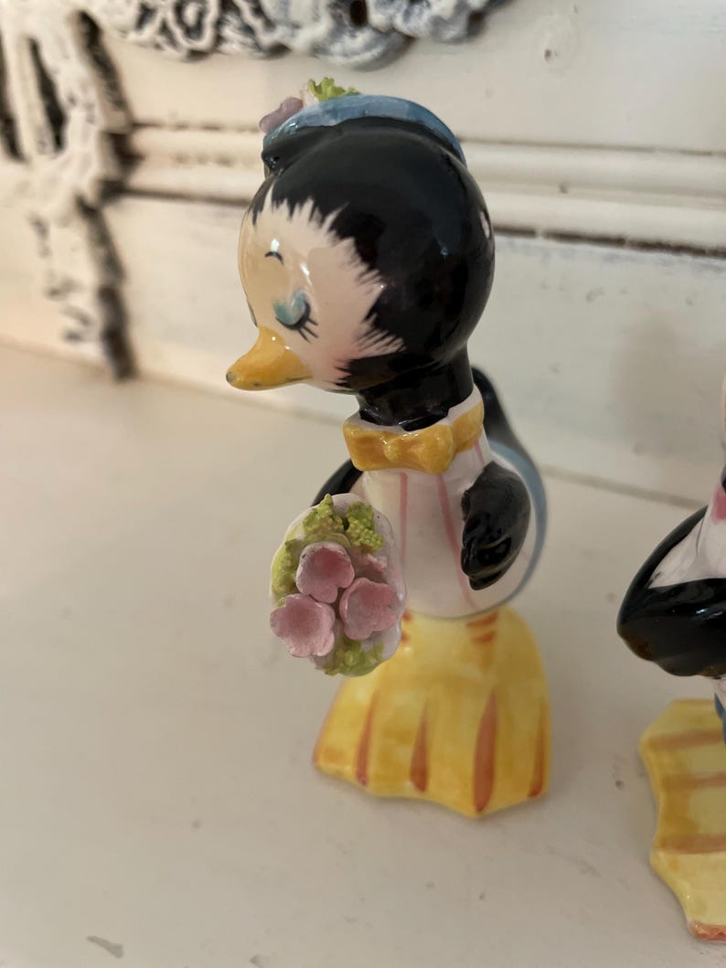 May include: A pair of ceramic penguin salt and pepper shakers. The female penguin is wearing a blue hat and holding a bouquet of pink flowers. The male penguin is wearing a black bow tie.