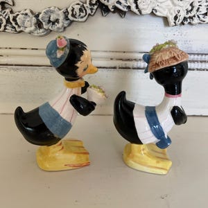 May include: A pair of ceramic salt and pepper shakers shaped like ducks. The duck on the left is wearing a blue hat with pink flowers and a blue and white striped shirt. The duck on the right is wearing a brown hat with flowers and a blue and white striped shirt. Both ducks have black bodies and yellow feet.