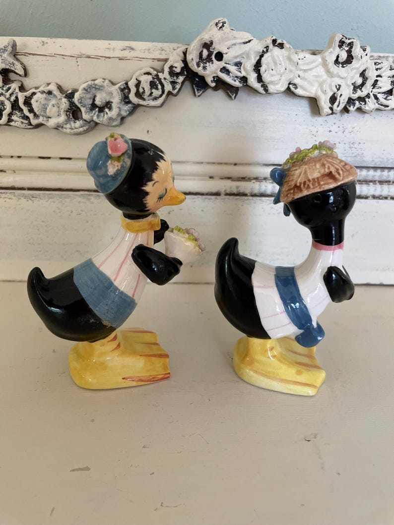 May include: A pair of ceramic salt and pepper shakers shaped like ducks. The duck on the left is wearing a blue hat with pink flowers and a blue and white striped shirt. The duck on the right is wearing a brown hat with flowers and a blue and white striped shirt. Both ducks have black bodies and yellow feet.