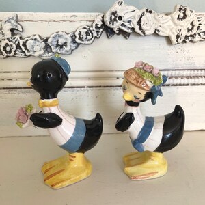May include: A pair of vintage ceramic duck salt and pepper shakers. The male duck is holding a bouquet of pink flowers and the female duck is wearing a hat with pink flowers. Both ducks are wearing blue and white striped shirts and have black and white bodies.