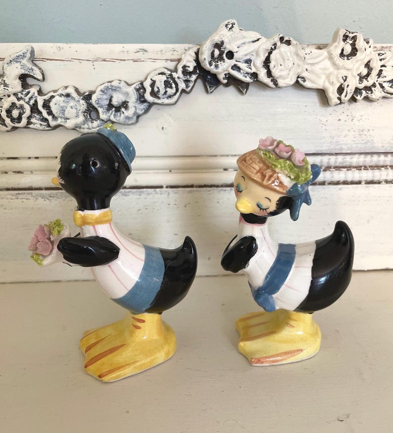 May include: A pair of vintage ceramic duck salt and pepper shakers. The male duck is holding a bouquet of pink flowers and the female duck is wearing a hat with pink flowers. Both ducks are wearing blue and white striped shirts and have black and white bodies.