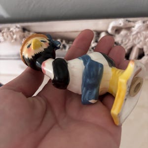 May include: A ceramic figurine of a bird wearing a blue and yellow hat and a blue and white dress. The bird is perched on a yellow and white base.