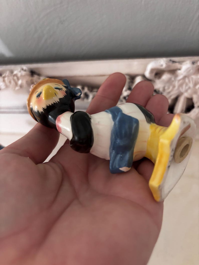 May include: A ceramic figurine of a bird wearing a blue and yellow hat and a blue and white dress. The bird is perched on a yellow and white base.