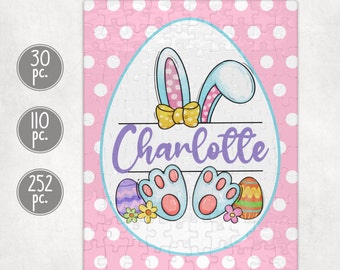 May include: A pink and white jigsaw puzzle with a white oval in the center. The oval has a cartoon bunny with blue ears and yellow bow, two blue bunny feet, and three Easter eggs. The name "Charlotte" is written in purple letters on the oval.