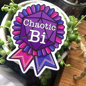 May include: A sticker with a purple, geometric design and the words "Chaotic Bi" in white. The sticker has a ribbon design with pink and blue accents. The sticker is placed on a small white pot with a plant.