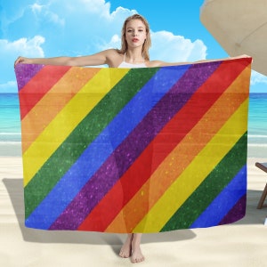 May include: A rainbow-colored sarong with a glittery texture. The sarong is draped over a person's shoulders and is being held in place by their hands.