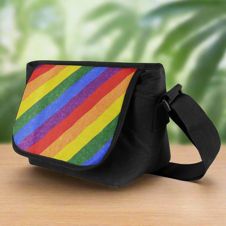 Puede incluir: Una bolsa de mensajero negra con una solapa de tela a rayas de arco&iacute;ris. Las rayas son diagonales y tienen una textura brillante.
