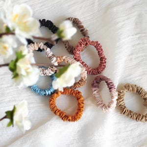 May include: A collection of ten circular fabric hair scrunchies in various colors including black, brown, dusty rose, light blue, mustard yellow, and beige. Each scrunchie features a gathered, ruffled texture and a smooth, satin-like finish.