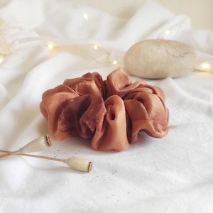 May include: A large, rust-colored scrunchie made of a silky material. The scrunchie is the focal point, resting on a white, textured surface. Dried seed pods and a small stone are also visible.