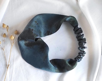 May include: A dark blue and teal tie-dye headband with a smooth, wide front section and a gathered elastic back band. The fabric features subtle variations in color intensity, creating a mottled appearance across the surface of the accessory.