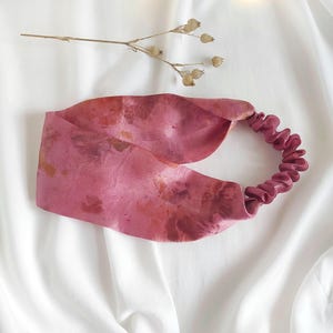 Könnte beinhalten: Ein breites Stirnband mit einem gesprenkelten Muster in Rosa und Dunkelrosa mit glatter Oberfläche. Das Accessoire verfügt über einen gerafften Gummizug auf der Rückseite für einen sicheren Sitz, begleitet von einem kleinen, getrockneten Zweig.