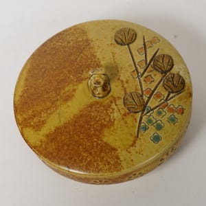 Puede incluir: Un recipiente redondo de cerámica con tapa. La tapa es de color beige con un patrón marrón texturizado y un diseño floral en marrón, naranja y verde. En la parte superior hay un pequeño botón redondo. El recipiente probablemente se utiliza para el almacenamiento.