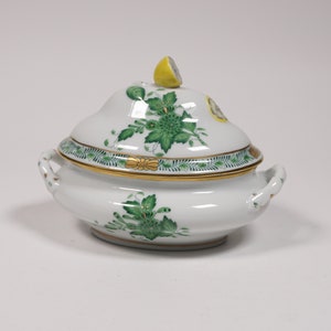 May include: A white porcelain serving dish with a green and gold floral design. The dish has a lid with a yellow lemon on top.