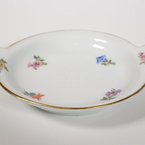 May include: A white porcelain dish with a gold rim and floral designs. The dish has two handles on the sides.