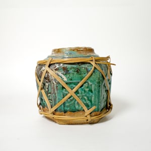 Peut inclure: Un petit pot en céramique hexagonale avec une finition émaillée vert et bleu sarcelle vif. La surface présente des motifs en relief et est entourée d'un treillis décoratif composé de fines bandes de bambou brun clair.