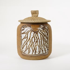 Puede incluir: Un tarro de cerámica hecho a mano con tapa. El tarro es de color marrón cálido con una tapa texturizada y un diseño blanco y marrón en la parte delantera. El diseño se asemeja a raíces o ramas.