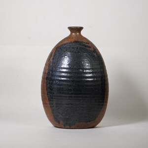 May include: A handmade ceramic vase with a dark brown and black glaze. The vase has a rounded body with a narrow neck and a textured surface. The dark glaze covers most of the vase, with a brown band around the top and bottom.