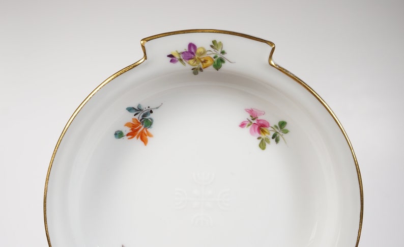 May include: A white porcelain bowl with a gold rim and hand-painted floral designs. The bowl has a scalloped edge and a small handle on each side.