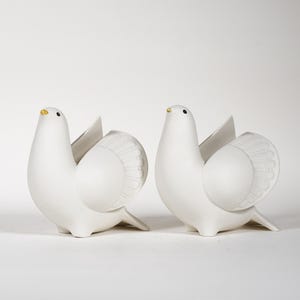 May include: Two white ceramic dove figurines with detailed wing designs. Each dove has a small, yellow beak and black eyes. The figurines are positioned side-by-side, showcasing their elegant, minimalist design. Ideal for home decor.