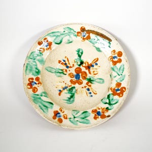 May include: A hand-painted ceramic plate with a cream base. It features a floral design with orange, blue, and green accents. The rim is decorated with green leaves and orange flowers. A unique piece for serving or display, approximately 20 cm in diameter.