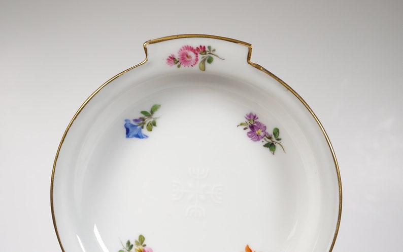 May include: A white porcelain dish with a gold rim and floral decorations. The dish has a scalloped edge and a small handle on one side.