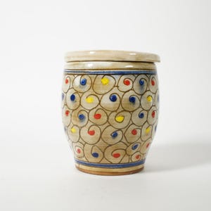 May include: A ceramic jar with a lid, featuring a beige base color and a pattern of swirling brown lines with red, yellow, and blue dots. The jar has a blue band near the base and top.