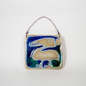 Op de afbeelding: Een vierkante keramische wandplaat met een glanzende blauwe en groene abstracte vogelachtige figuur op een gespikkelde beige achtergrond. Het object hangt aan een dunne, donkerbruine metalen draad bevestigd aan twee kleine lussen.