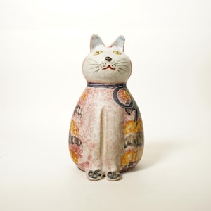 May include: A ceramic cat figurine with a seated pose. The cat has a white face with yellow eyes, a red mouth, and a blue collar. The body is primarily pink with orange and yellow patterns. The cat's ears have blue tips.