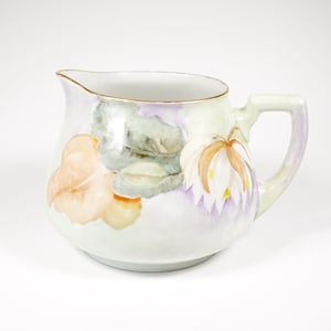 May include: A white porcelain creamer with a floral design. The creamer has a rounded body with a handle and a gold rim. The flowers are painted in shades of pink, yellow, and green.