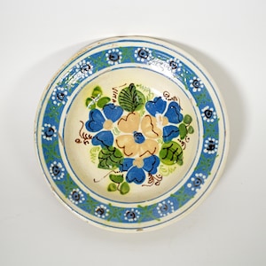 May include: A round, hand-painted ceramic plate with a floral design. The plate features a cream-colored center with blue and yellow flowers, surrounded by a blue and green border with white accents. The plate has a rustic, vintage aesthetic.