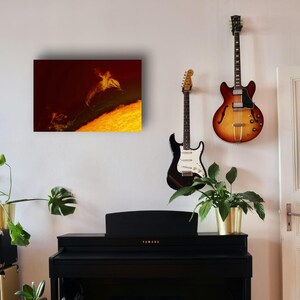 May include: A black upright piano sits below a rectangular wall art piece featuring a golden dolphin silhouette against a dark red background. To the right, a black electric guitar and a sunburst hollow-body electric guitar hang on the wall.