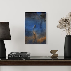 May include: A dark blue and orange abstract art print with a vertical, cloud-like shape in the center. The print is hanging on a white wall above a dark brown wooden table with a black lamp and a vase with dried flowers.