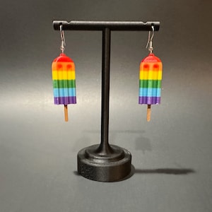 May include: Rainbow popsicle dangle earrings. The earrings are made of polymer clay and feature a rainbow design with a purple, blue, green, yellow, orange, and red color scheme. The earrings are hanging from silver hooks.
