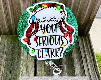 Puede incluir: Un carrete de insignia redondo y verde brillante con la frase "YOU SERIOUS CLARK?" en texto negro. El diseño presenta un gorro de Papá Noel con luces navideñas. La parte inferior del carrete de la insignia tiene un clip plateado y el texto "20 COLOR OPTIONS".