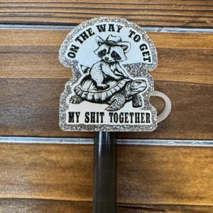 May include: A pen topper with a raccoon wearing a cowboy hat riding a turtle. The pen topper has the text "ON THE WAY TO GET" and "MY SHIT TOGETHER" in a glittery frame. The pen is black.